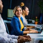 Three diverse professionals working and smiling at office desks, fostering teamwork and collaboration.